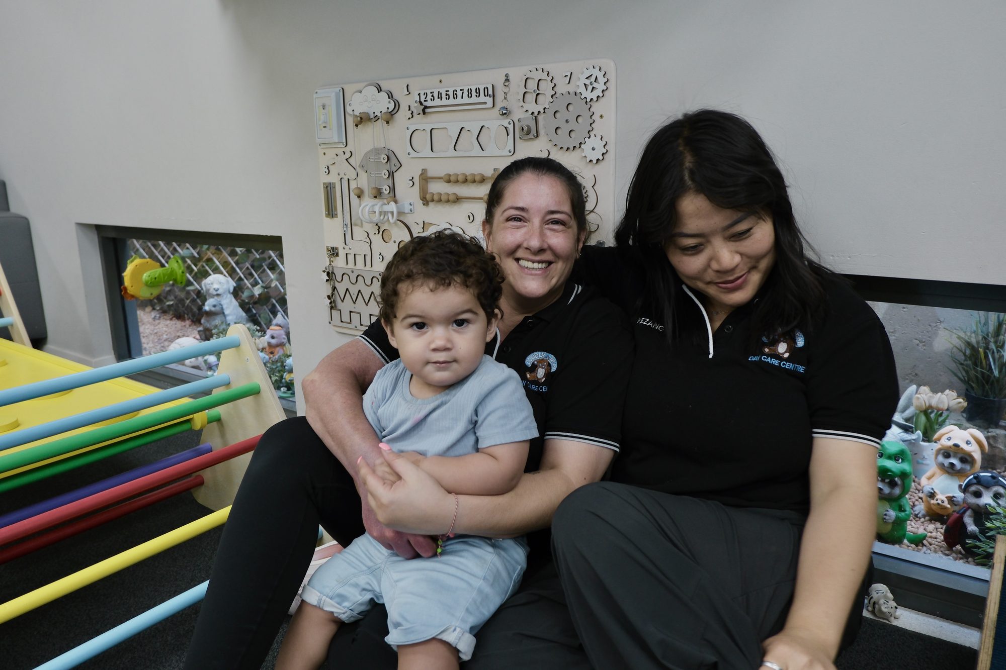 Two educators sitting with a toddler in front of a wall activity board